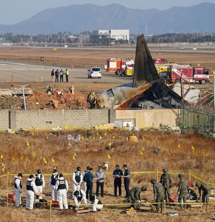 muan-airport-crash-site-investigation.jpg 무안공항 활주로와 소방차들을 배경으로 처참하게 부서진 여객기 꼬리 날개 잔해가 보이고 노란색 표지 깃발이 가득 꽂힌 풀밭 통제선 안에서 군인과 경찰 요원들이 유해 및 잔해를 수색하고 있는 다큐멘터리 보도사진